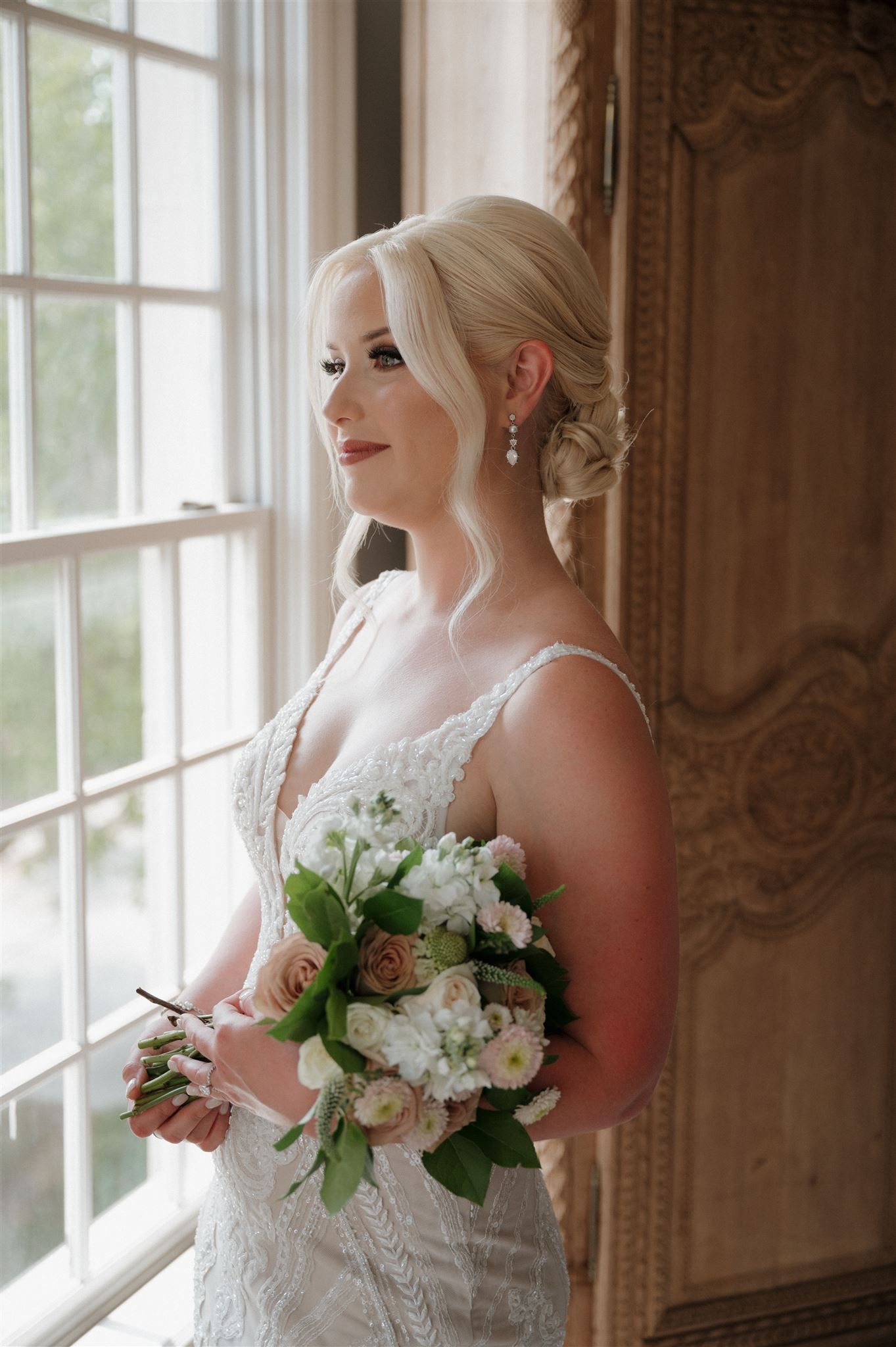 Elegant bridal portrait by a window featuring a white lace gown and fresh floral bouquet.