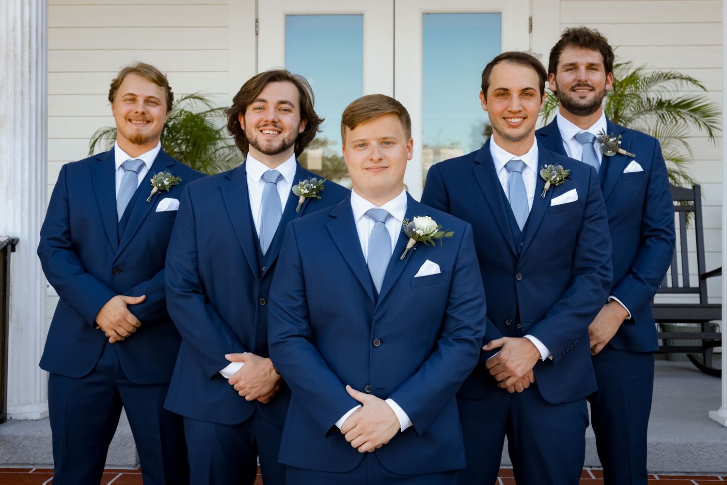 Groomsmen in matching navy suits and light blue ties pose on a white porch.