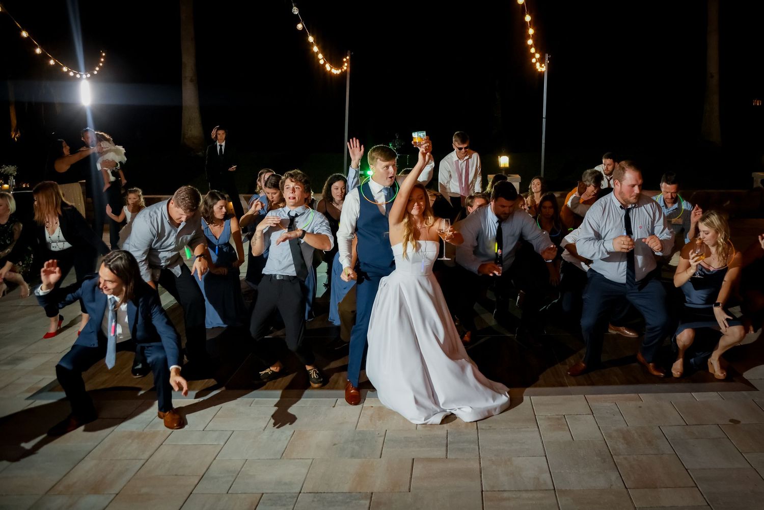 Wedding party celebrates with energetic dance moves on an outdoor patio under evening lights.