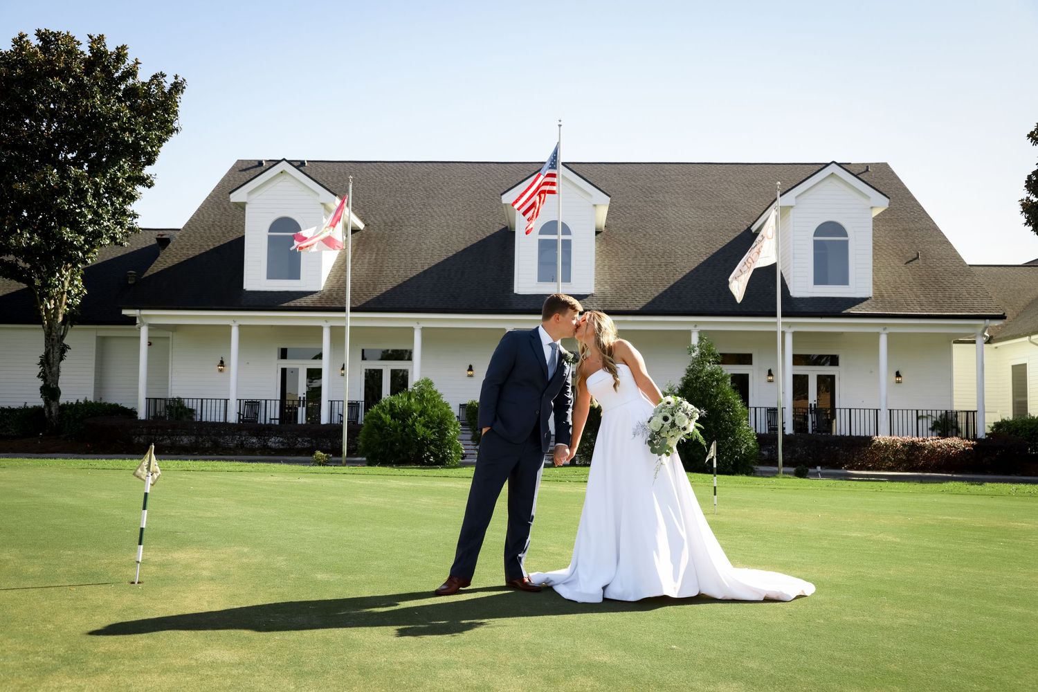 A newlywed couple poses on a golf course with a classic white clubhouse in the background.