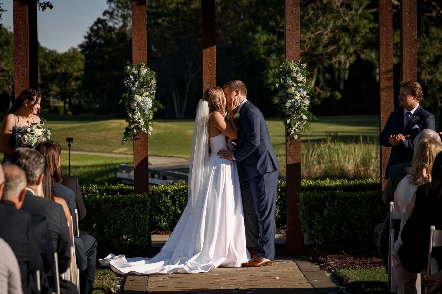 An outdoor wedding ceremony under wooden pillars with floral arrangements and seated guests.