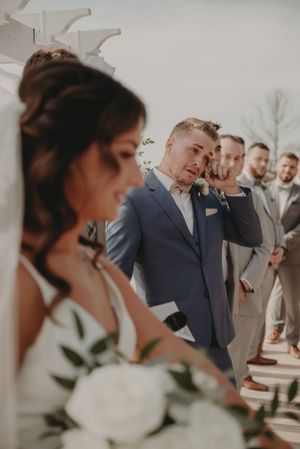 Members of the wedding party share emotional reactions during an outdoor ceremony with white floral bouquets.