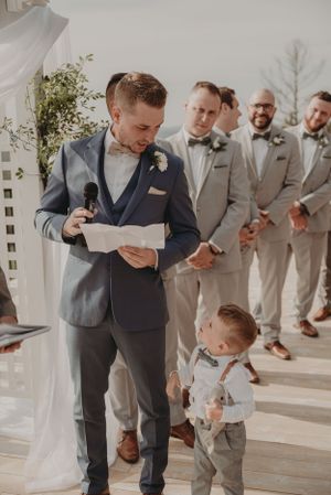 A touching moment during wedding vows on a sunlit terrace with groomsmen in the background.