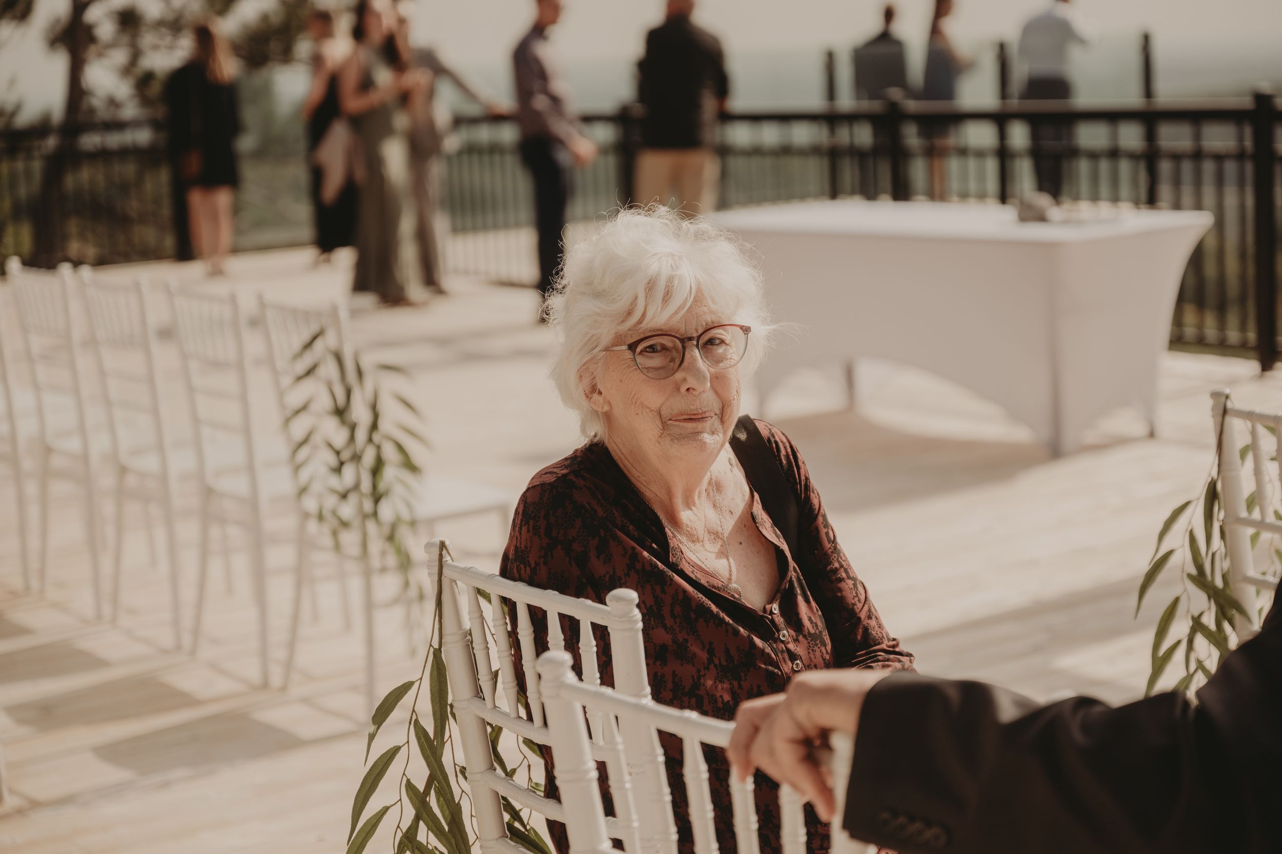 An elderly guest sits in a white chair at an outdoor wedding venue.