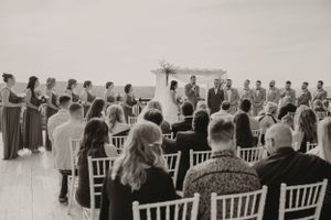 Black and white photo of outdoor wedding ceremony with guests seated in white chairs.