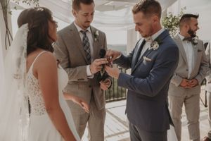 Wedding ceremony participants gather during ring exchange at outdoor venue with white drapery.
