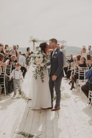A wedding ceremony taking place on an outdoor deck with guests seated in white chairs and a lush floral bouquet.