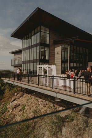 Modern wooden and glass building perched on a hillside with guests gathered on the outdoor deck.