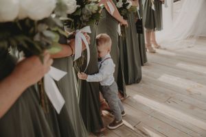 Small child in formal attire stands among long dresses during wedding ceremony.