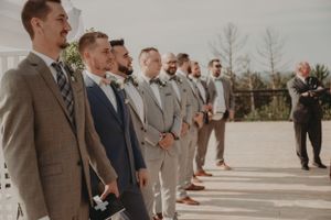 A line of groomsmen in coordinated formal attire stand during an outdoor ceremony.