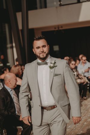 Groomsman in light gray suit and bowtie stands during wedding ceremony.