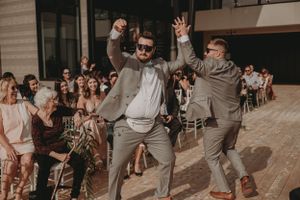 Wedding party members in grey suits dance enthusiastically at an indoor reception.