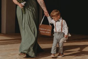 A small child in suspenders walks holding a rustic wooden sign.