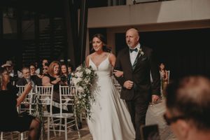 A couple walks down the aisle at indoor wedding ceremony with white chairs.
