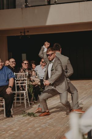 Wedding guest in gray suit dances enthusiastically on wooden floor during reception.