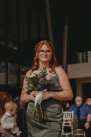 A bridesmaid in sage green dress holds a floral bouquet at an indoor wedding ceremony.