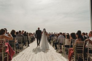 A couple walks down outdoor white aisle surrounded by guests in white chairs.