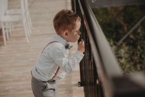 Young child in suspenders and dress shirt peers through outdoor railing during wedding event.