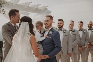 A wedding ceremony moment under a white pergola with groomsmen in grey suits looking on.