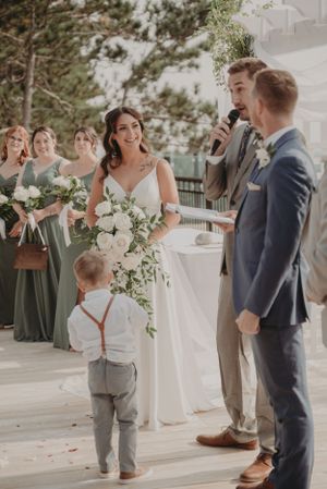 A small child in suspenders stands near adults during outdoor wedding ceremony with white floral arrangements.