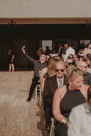 Wedding guests seated in rows during an indoor ceremony with one person standing enthusiastically.