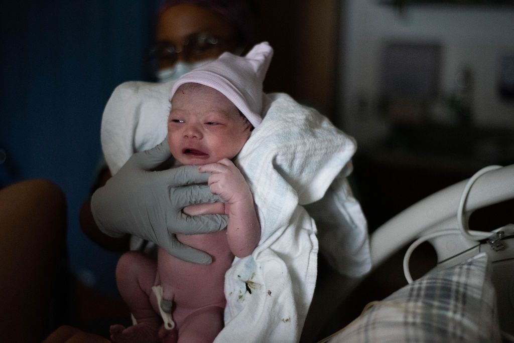 Healthcare worker holding a crying newborn baby wrapped in white blanket.