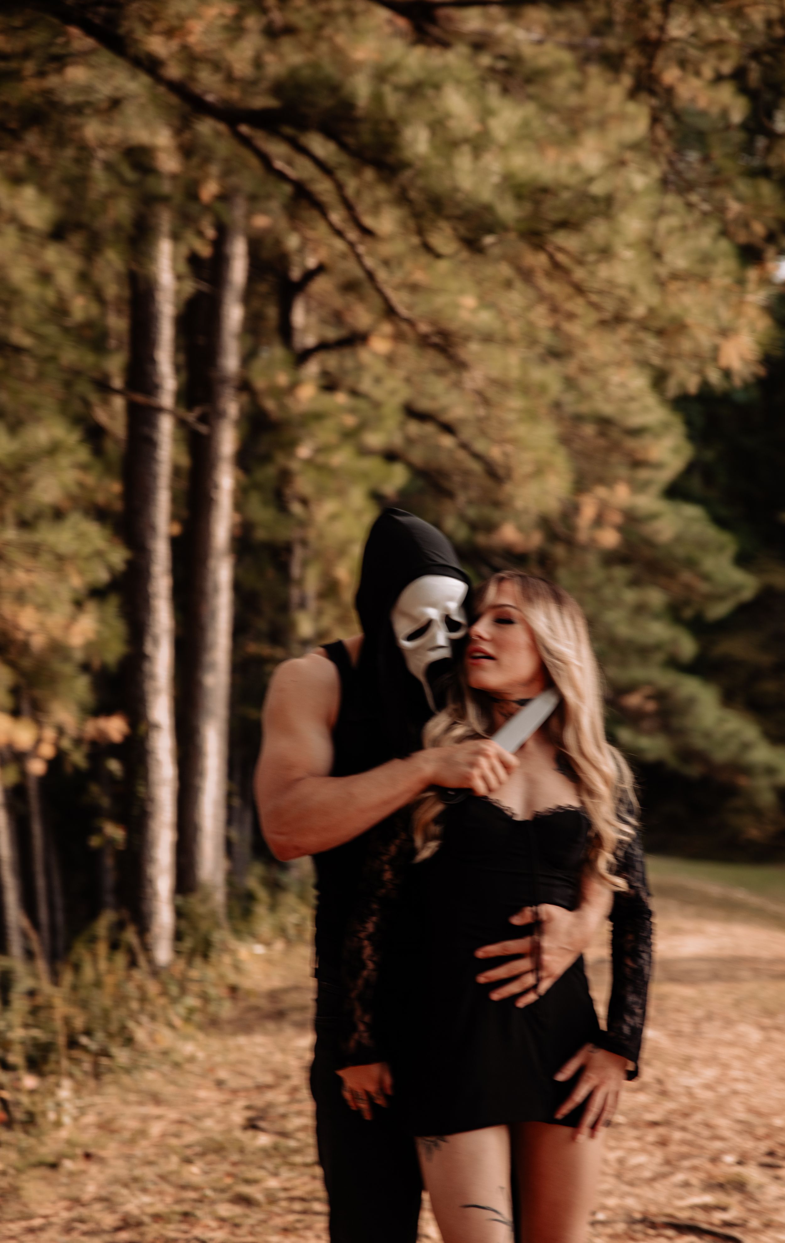 A couple in black clothing embraces on a sunlit forest path.