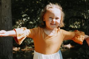 Young child twirls joyfully in a colorful tiered dress against a wooded backdrop.