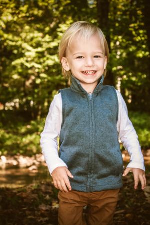 Small child wearing a blue vest and brown pants smiles warmly in an autumn forest setting.