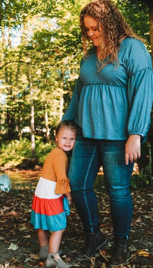 Parent and child share a tender moment while wearing coordinating teal and orange outfits in a sunlit forest setting.