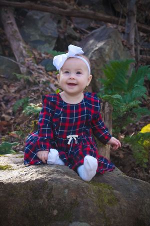 Baby in plaid holiday dress and white bow sits smiling on mossy rock in woodland setting.