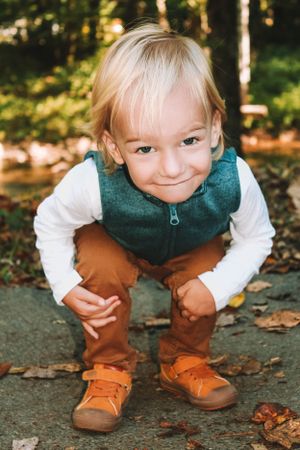Child in teal vest and brown pants crouches playfully on a leaf-covered path.