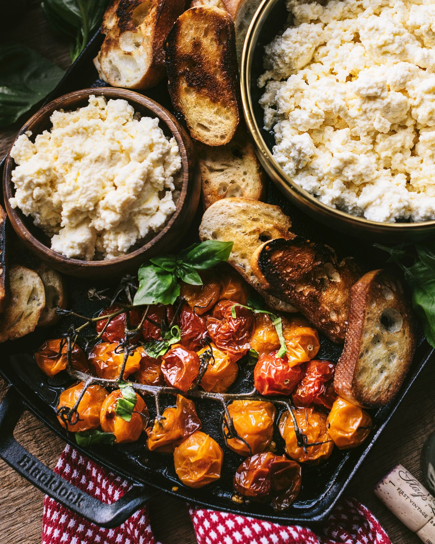 Close-up of roasted cherry tomatoes in a cast iron skillet alongside bowls of fluffy ricotta and toasted bread.