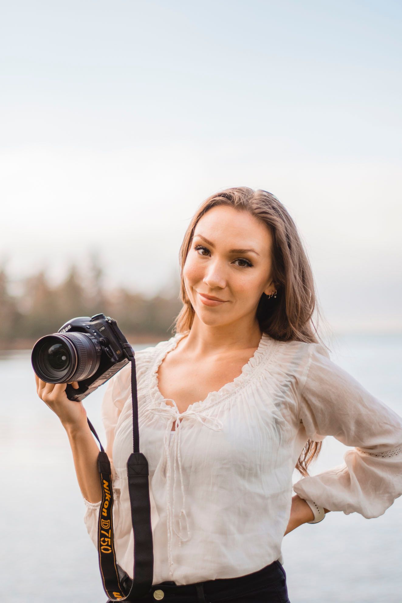 Professional photographer in a white blouse holds a DSLR camera near a lake at sunset.