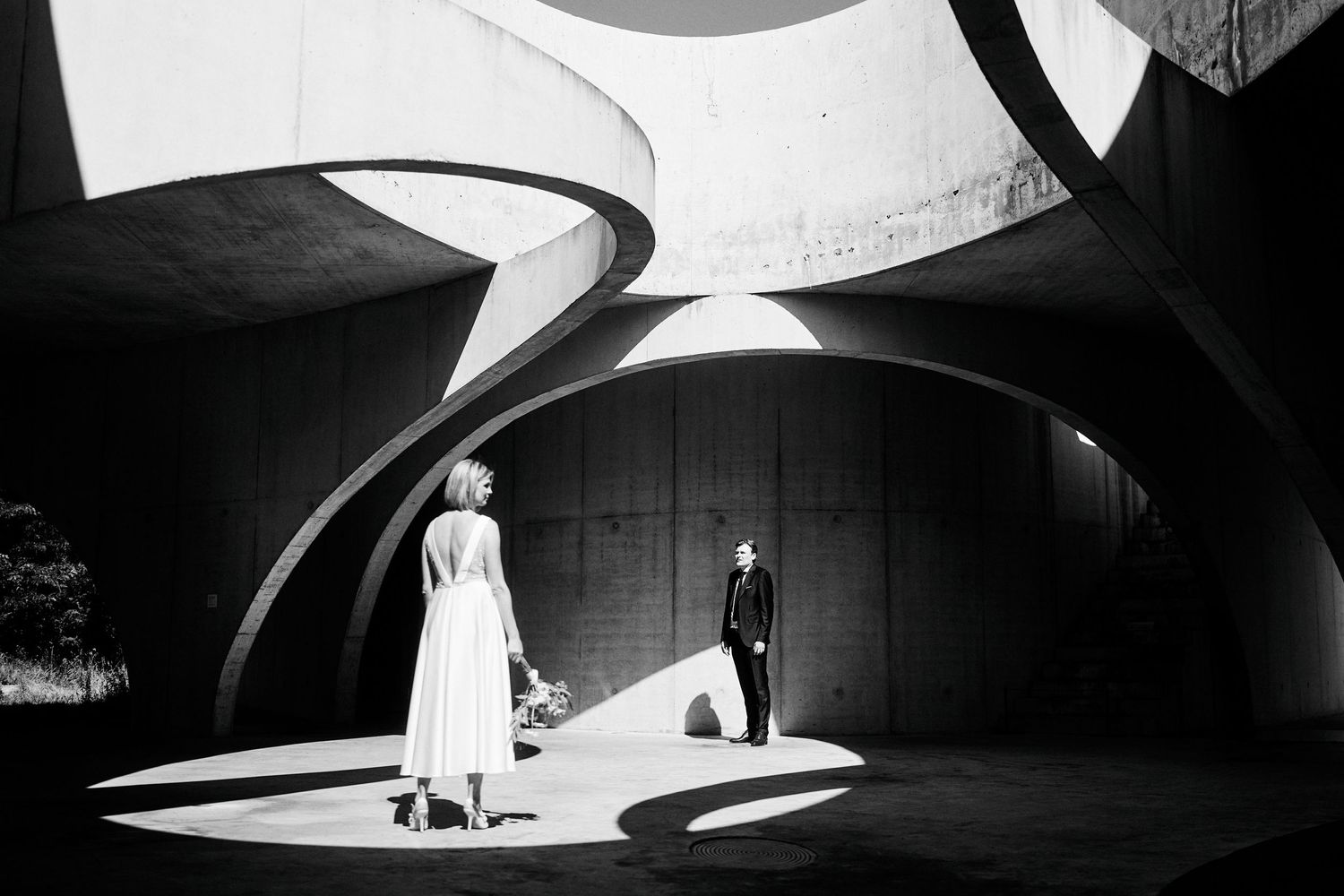 Een dramatische zwart-wit theaterscène met gebogen architectonische elementen en een grimmige schijnwerper die twee figuren verlicht.