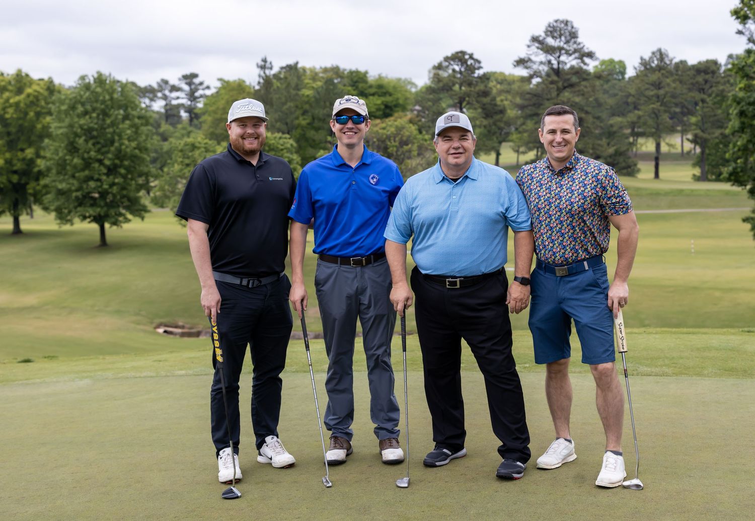Group of golfers pose together on green of golf course with trees in background.