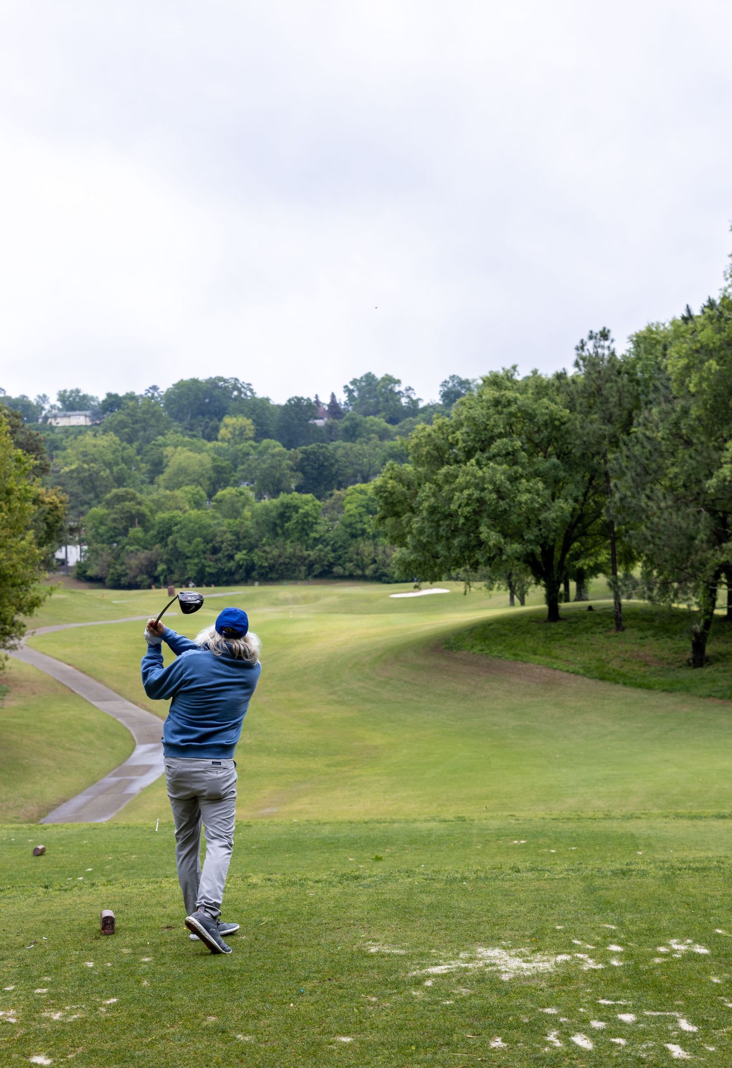 Golfer mid-swing on scenic hillside course with trees and rolling landscape.