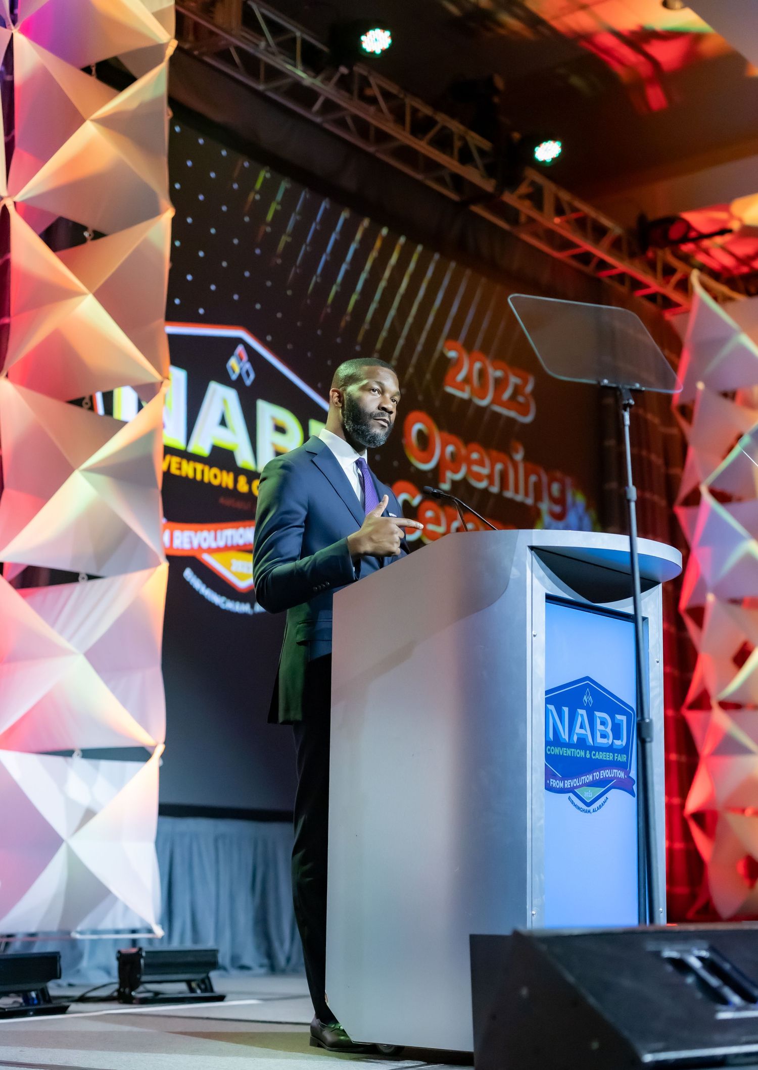 Speaker presents at NABJ opening ceremony from behind podium on stage with geometric backdrop lighting.