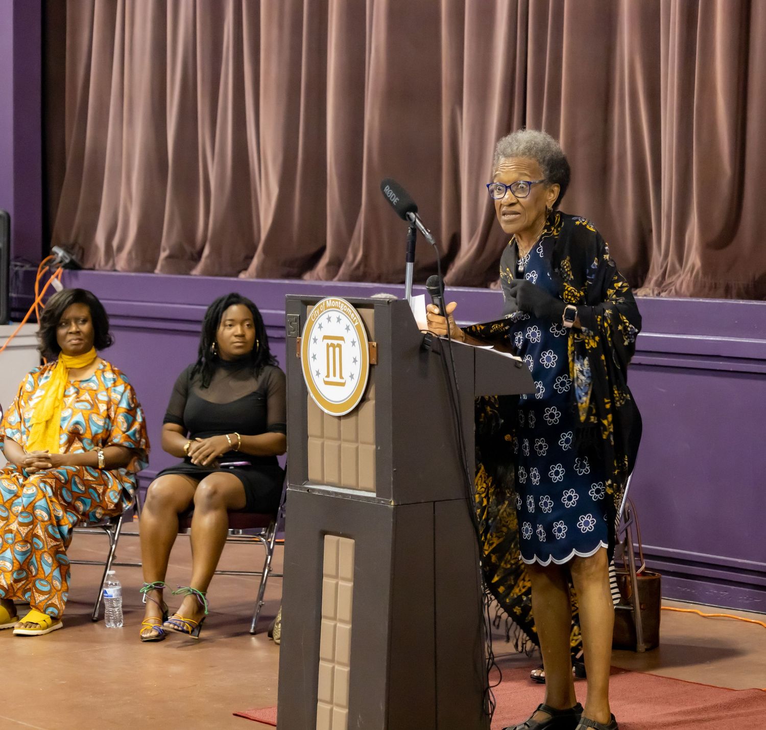 Speaker addresses audience from wooden podium on stage with purple curtain backdrop.