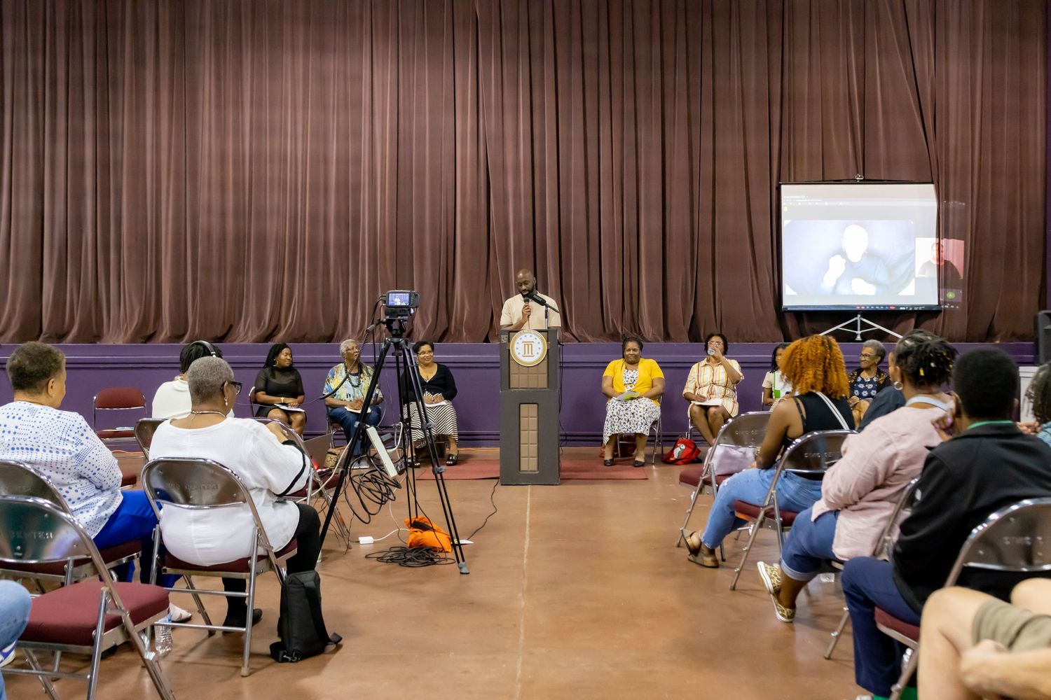 Audience members seated in folding chairs watch presentation in conference room with stage and projector screen.