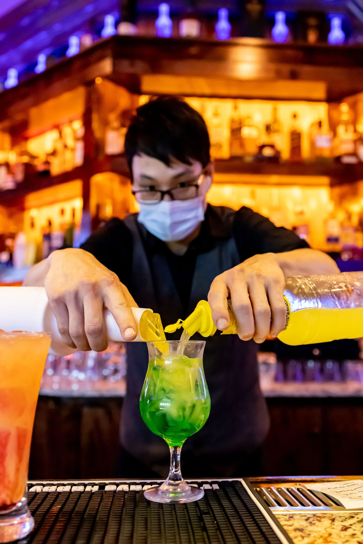 Bartender making a vibrant green cocktail at an illuminated bar counter.