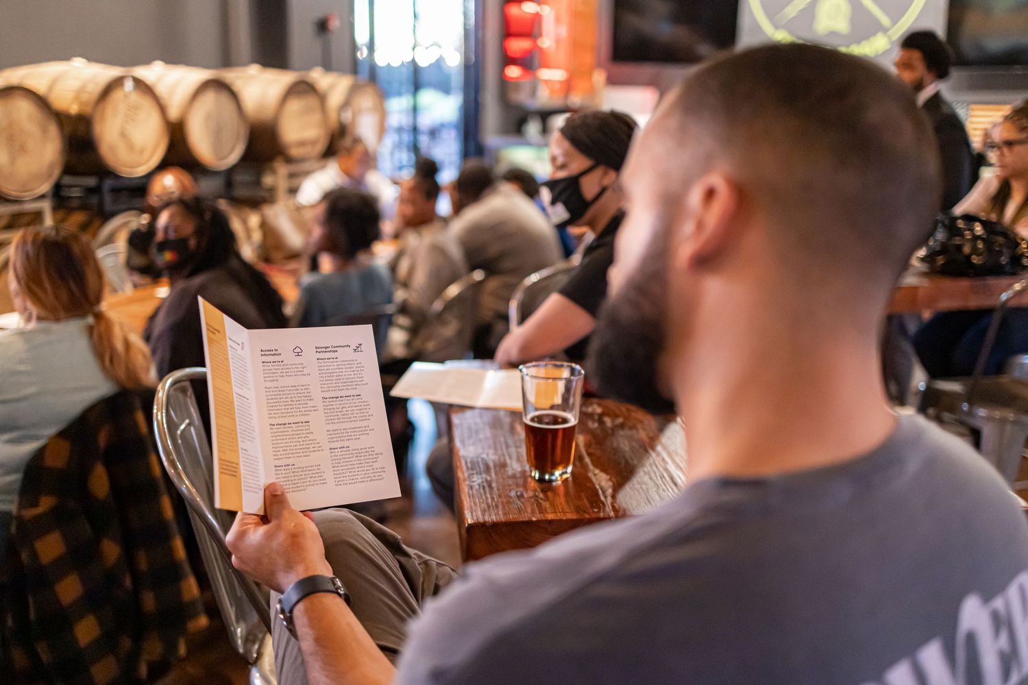 Customer reads menu while enjoying a drink in a rustic brewery setting with wooden barrels in background.