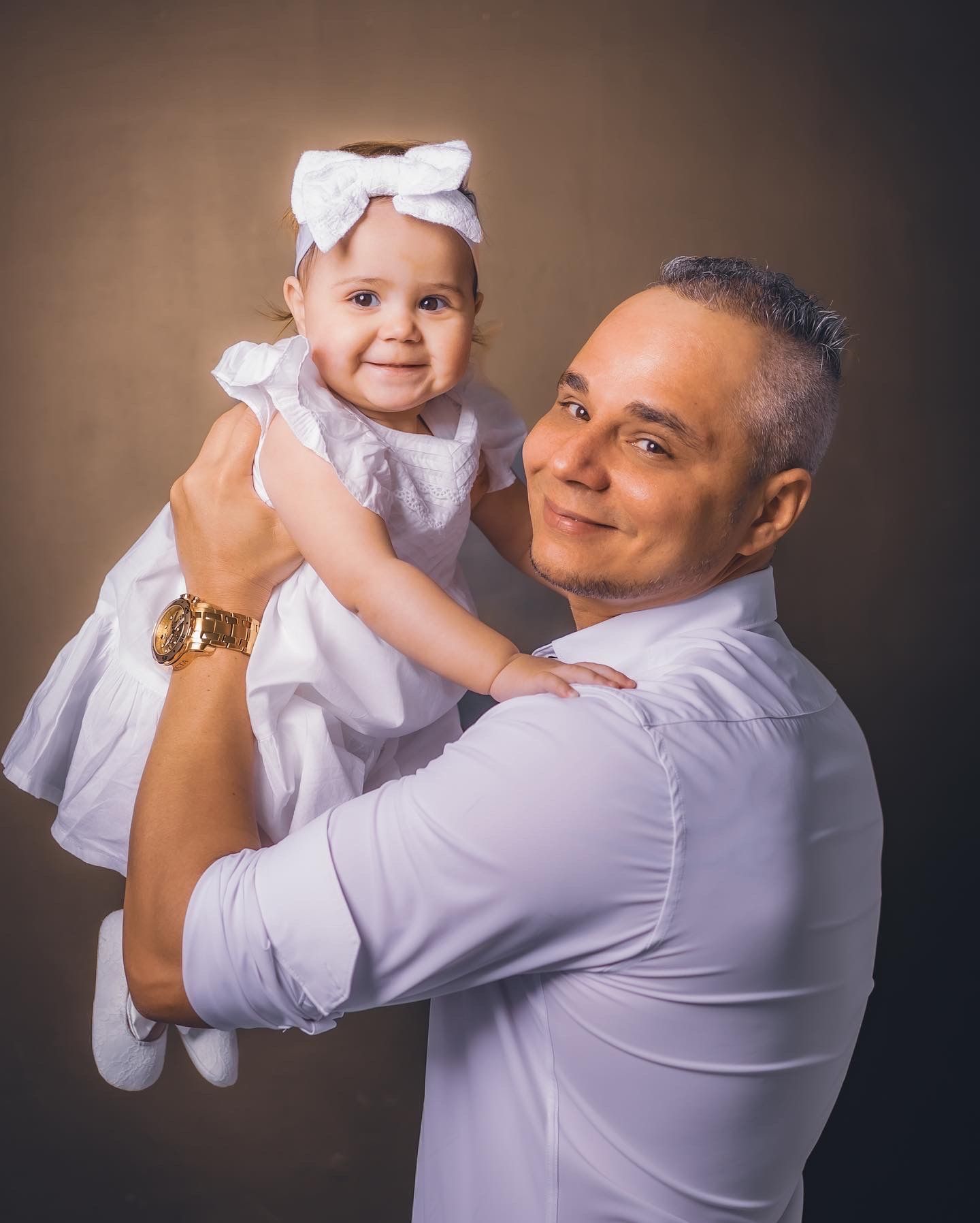 Proud father smiles while holding his baby daughter dressed in white with a bow headband.