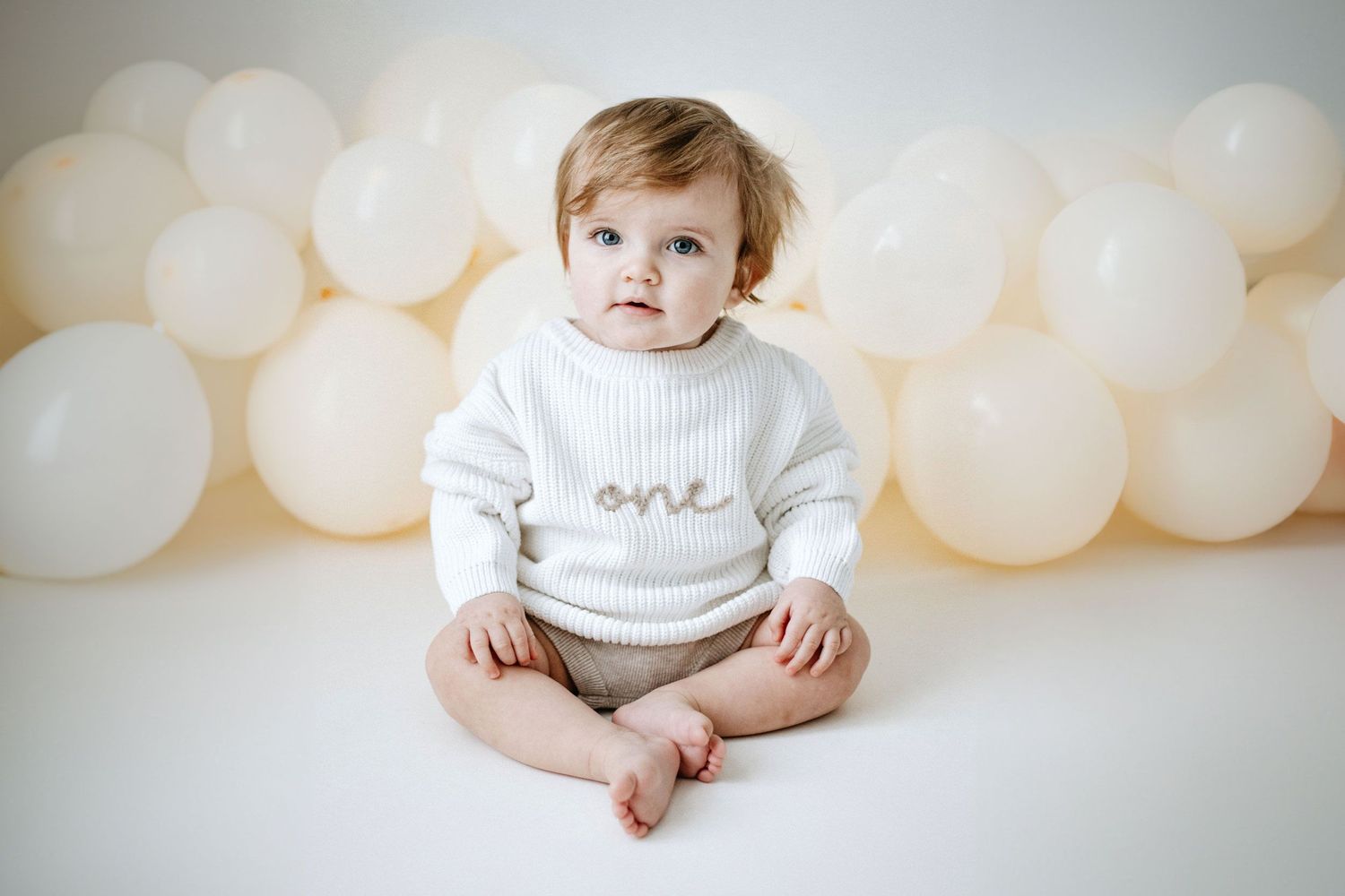 Baby wearing white knit sweater with 'one' text sits against white backdrop with soft balloon decorations.