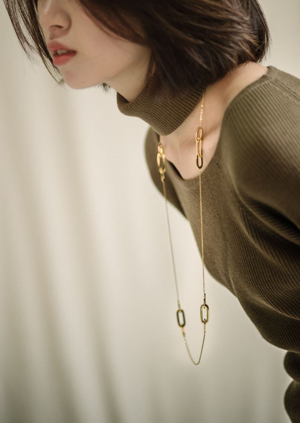 Elegant fashion detail of long necklace and olive sweater in soft lighting.
