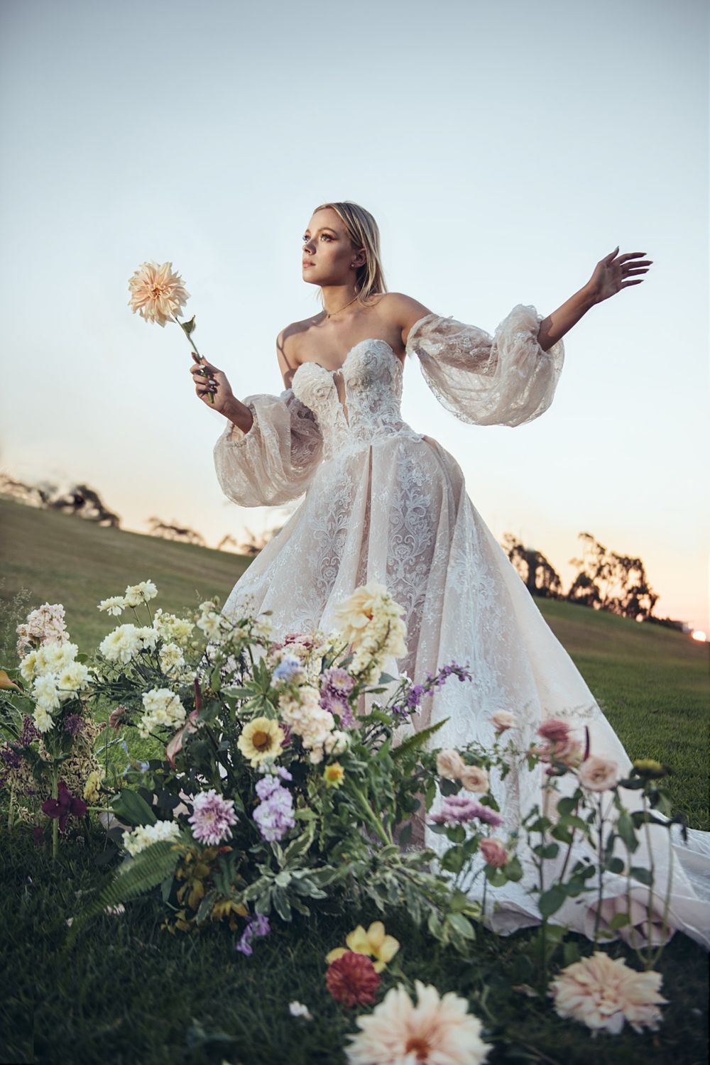 A dreamy wedding portrait with a lace gown and flowing sleeves amid garden flowers at dusk.