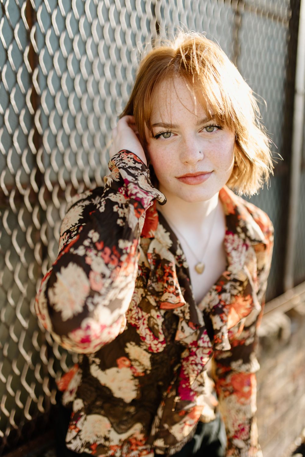 red haired high school senior in a downtown setting. she is sitting on low ledge with rusted cyclone fence behind her.