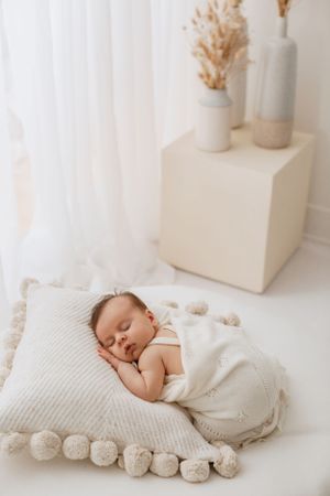 Sleeping baby in white clothing rests on decorative pillow with pom pom trim in minimalist nursery setting.