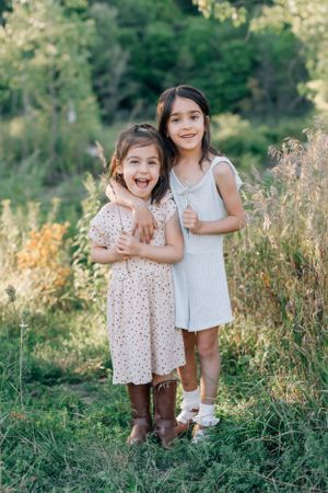 Two children in dresses pose together outdoors in a field of tall grass.