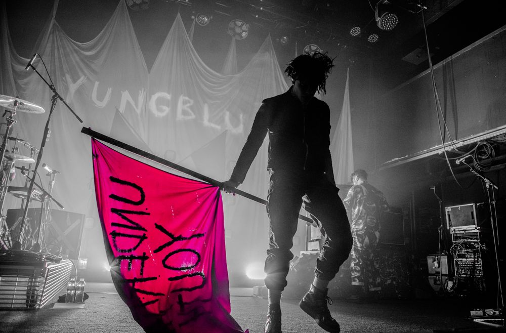 A silhouetted performer holds a bright pink flag on a dimly lit concert stage with dramatic backlighting and fog effects. Youngblud